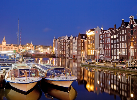 City of Amsterdam in Netherlands at night, historic apartment houses with reflections on water and boats ready for canal tours and cruises.