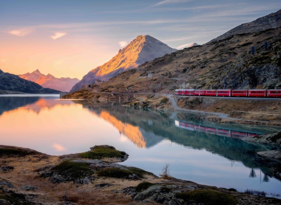 Aerial view of Bernina Express train near Bianco lake in the Swiss Alps at sunset, with glowing mountain peaks, water with reflections, rocky shore. Peaceful autumn landscape. Mountain train in fall
