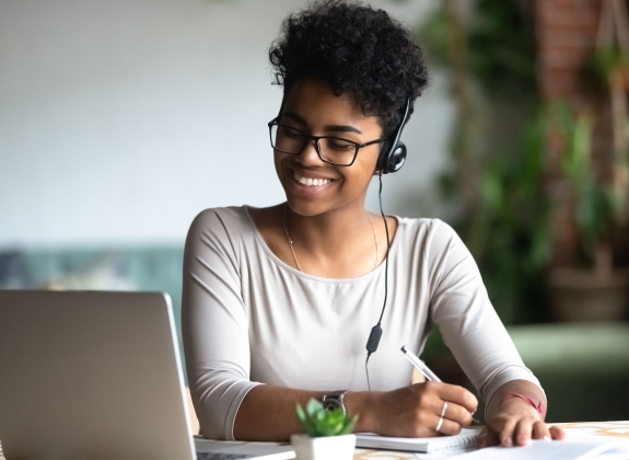 Smiling millennial female student in headphones and glasses sit at desk watch webinar making notes, happy biracial young woman in earphones work study using computer