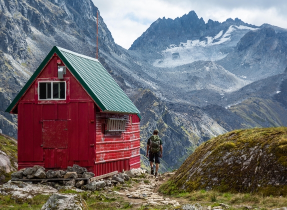 A man with a backpack out hiking walks up the trail to a red and green wilderness hut deep in the Talkeetna Mountains near Hatcher Pass, Alaska