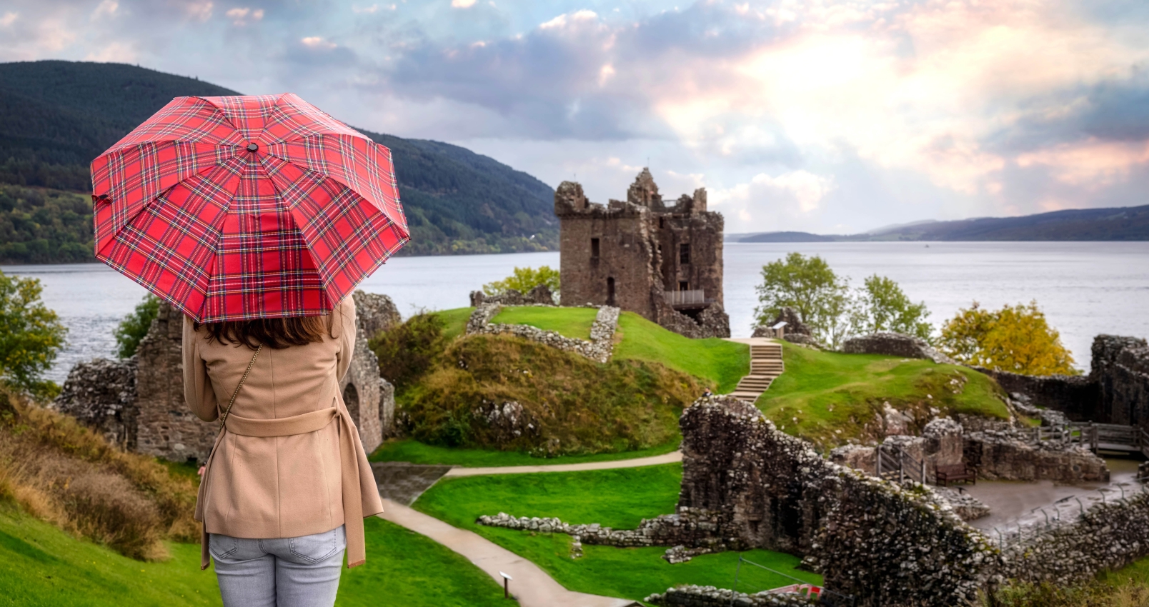 A tourist woman with a scottisch pattern umbrella looks at the famous Urquhart Castle at Loch Ness during autumn time, Scotland