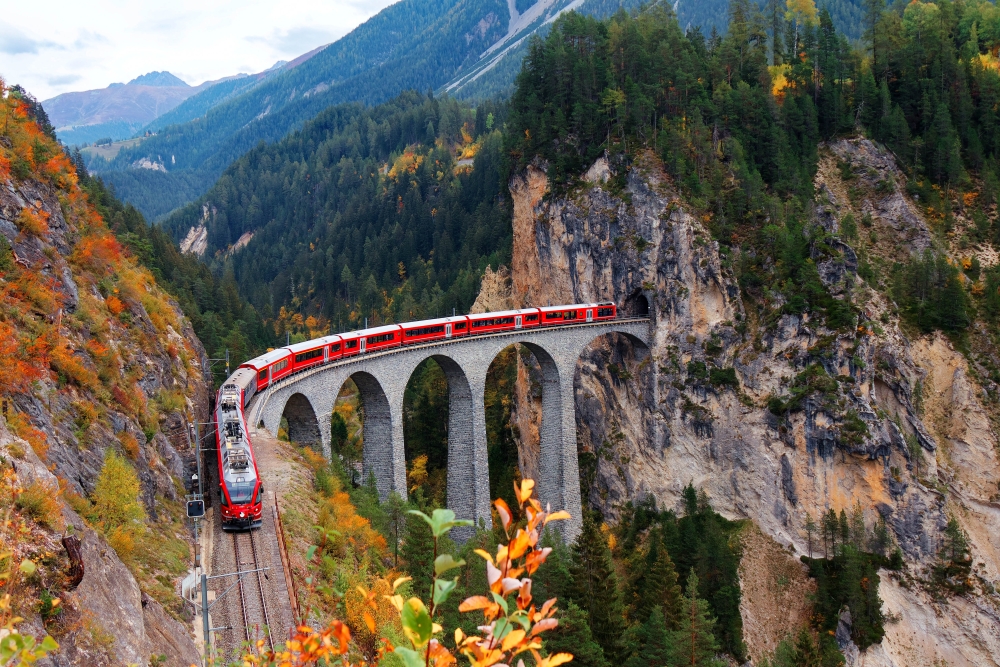 Glacier Express train coming out of the tunnel in a cliff crossing famous Landwasser Viaduct over a deep gorge with fall colors on the rocky mountainside in Filisur, Grisons, Switzerland